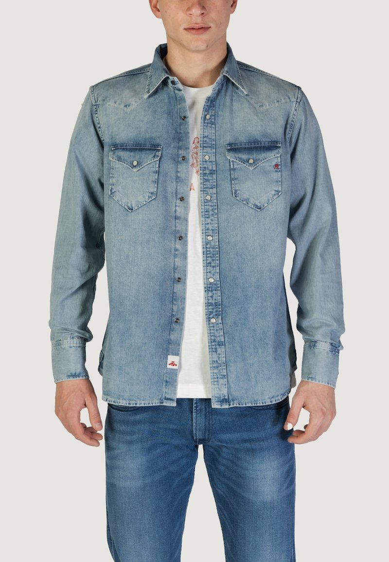 Camicia di jeans azzurro chiaro con due taschini sul petto, colletto a punta e bottoni metallici. Indossata sopra una t-shirt bianca e abbinata a jeans scuri.