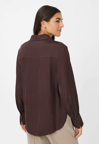 Camicia lunga in raso marrone con colletto, polsini con bottone e orlo curvato. Texture liscia, vestibilità comoda e dettaglio di cucitura sul retro discreto.