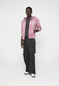 Ανοιχτό ροζ φούτερ με το "GAP" σε κρέμα, γκρι μπλουζάκι από κάτω, σκούρες cargo παντελόνες και γκρι αθλητικά παπούτσια με λευκές λεπτομέρειες. Απλή φόντο.