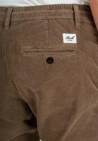 Brown corduroy pants feature a back pocket with a black button and a white label with "Reell" text. The fabric has a soft texture and ribbed pattern.