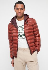 Chaqueta acolchada roja con un acabado brillante, capucha y puños de canalé. Lleva puesta sobre una camiseta gráfica blanca y pantalones beiges.
