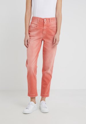 Personne portant un jean taille haute rose corail et des baskets blanches debout devant un fond clair uni.