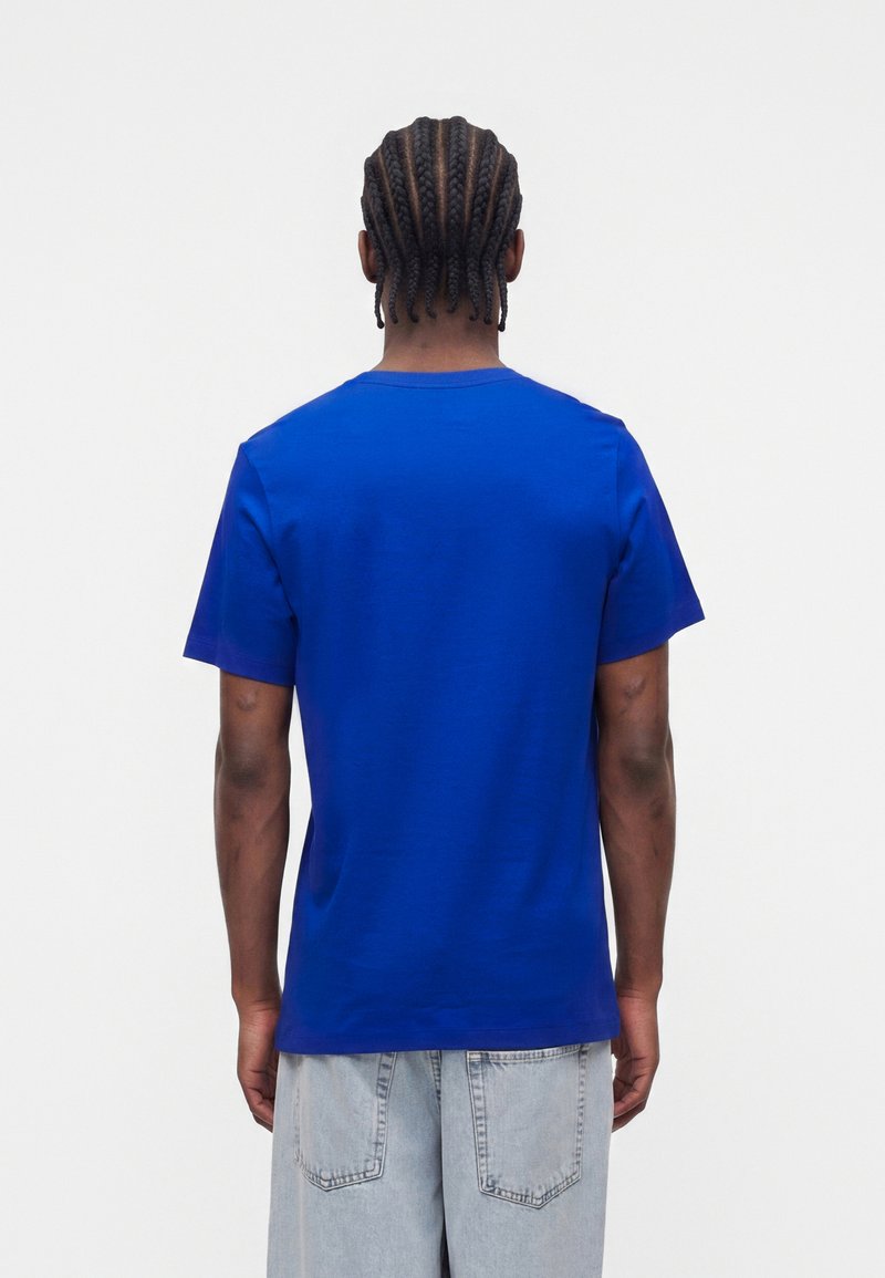 Person mit geflochtenen Haaren, die ein einfaches blaues Kurzarmshirt und hellblaue Jeans trägt, von hinten vor weißem Hintergrund.