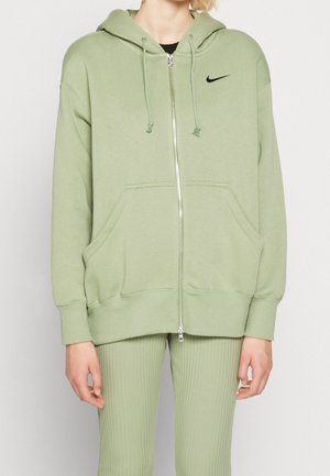 Person iført en lysegrøn Nike-hættetrøje med lynlås og sort logo samt matchende ribbede leggings, hænderne afslappet ved siderne, vendt fremad.