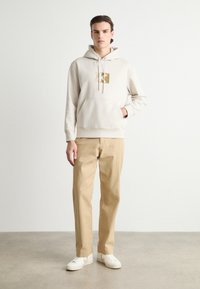 Hanorac bej cu un logo auriu pe piept, purtat cu pantaloni bej deschis și adidași albi. Materialul are o textură moale.