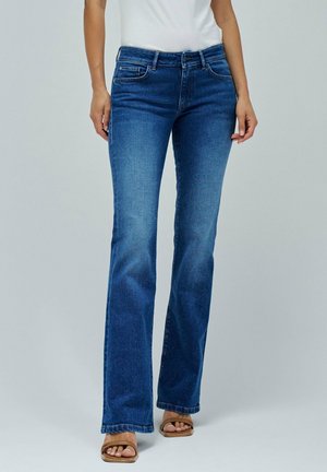 Kobieta w niebieskich dżinsach typu bootcut i beżowych sandałach z odkrytymi palcami stoi na jednolitym jasnym tle.