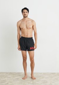 Juodi maudymosi šortai su elastingu juosmeniu, ant kairės kojos su raudonu "HUG" logotipu. Modelis be marškinėlių, stovi ant šviesios grindų.