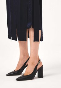 Tacchi slingback in pelle nera con punta affusolata, alto tacco a blocco e cinturino alla caviglia. La texture è liscia con una leggera finitura lucida.