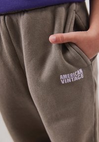 Close-up van een hand in de zak van olijfgroene sweatpants met "AMERICAN VINTAGE" in het wit gedrukt op het bovenbeen.