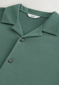 Polo shirt texturizado en verde con un collar suave, que presenta dos botones negros y una etiqueta etiquetada en el interior del collar. Diseño de ajuste regular.