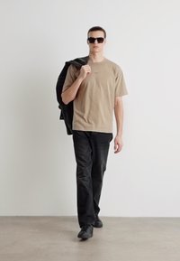 T-shirt marron clair, jeans foncés et chaussures noires. Le modèle porte une veste noire sur son épaule et des lunettes de soleil sombres. Fond uni.