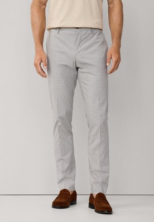 Man in een lichtgrijze slim-fit broek en bruine suède instappers, staand tegen een effen lichte achtergrond.