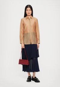Chemise boutonnée marron clair avec une texture légèrement transparente, associée à une jupe superposée bleu marine. Le mannequin tient un sac à main rectangulaire rouge.