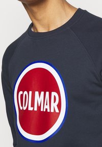 Marinblå sweatshirt med raglanärmar och en stor röd cirkulär logotyp med "COLMAR" i vit text med blå kant. Mjuk textur.