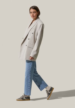 Helle beige, doppelt geknöpfte Jacke mit breitem Kragen, zwei Fronttaschen, kombiniert mit hellblauen, geraden Jeans und grauen Sneakers.