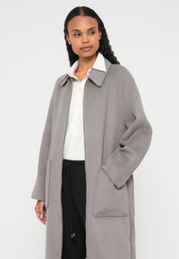 Harmaa villaseos-takki, jossa on jämäkkä kaulus, etutaskut ja rento istuvuus, kerrospukeutuneena valkoisen paidan ja mustien housujen päälle.