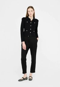 Schwarze taillierte Jacke mit goldenen Knöpfen und Schulterakzenten, kombiniert mit schwarzen, zulaufenden Hose und schwarzen, gemusterten Ballerinas. Schlichtes Design.