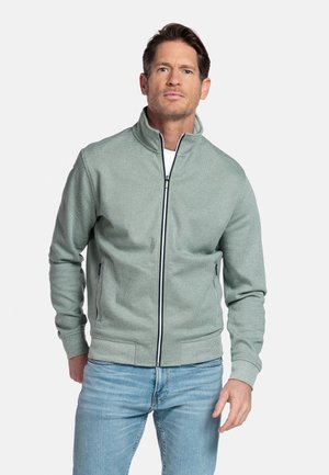 Homme portant une veste zippée vert clair et un jean bleu clair, debout devant un fond blanc uni.