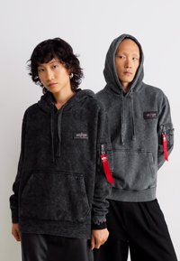 Zwei junge Erwachsene tragen dunkelgraue Kapuzenpullover mit Fronttaschen und roten Labels an den linken Ärmeln und posieren vor einem schlichten, hellen Hintergrund.