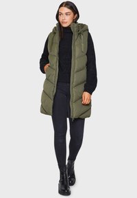 Gilet matelassé vert olive avec capuche, fermeture éclair à l'avant et quilting en chevrons ; porté sur un pull en maille noir et un pantalon noir slim avec des bottes à talons.