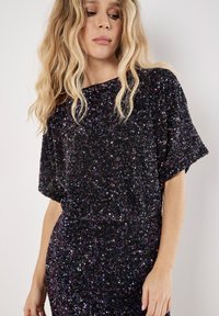 Schwarzes Pailletten-Top mit kurzen Ärmeln, das einen lockeren Schnitt und eine schimmernde Textur aufweist. Akzente aus lila und silbernen Pailletten überall.