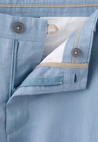 Pantalon bleu clair avec fermeture éclair à l’avant, deux boutons gris, passants beiges pour boutons, et bordure beige à l’intérieur de la taille et de la patte de la braguette.