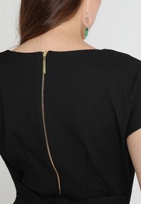 Femme portant une robe noire texturée avec une fermeture éclair dorée visible dans le dos et une longue boucle d'oreille pendante rose et verte.