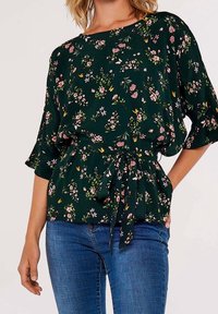 Blouse fleurie vert foncé à manches courtes avec un lien à la taille. Présente un mélange de petites fleurs roses et blanches ainsi que des feuilles.
