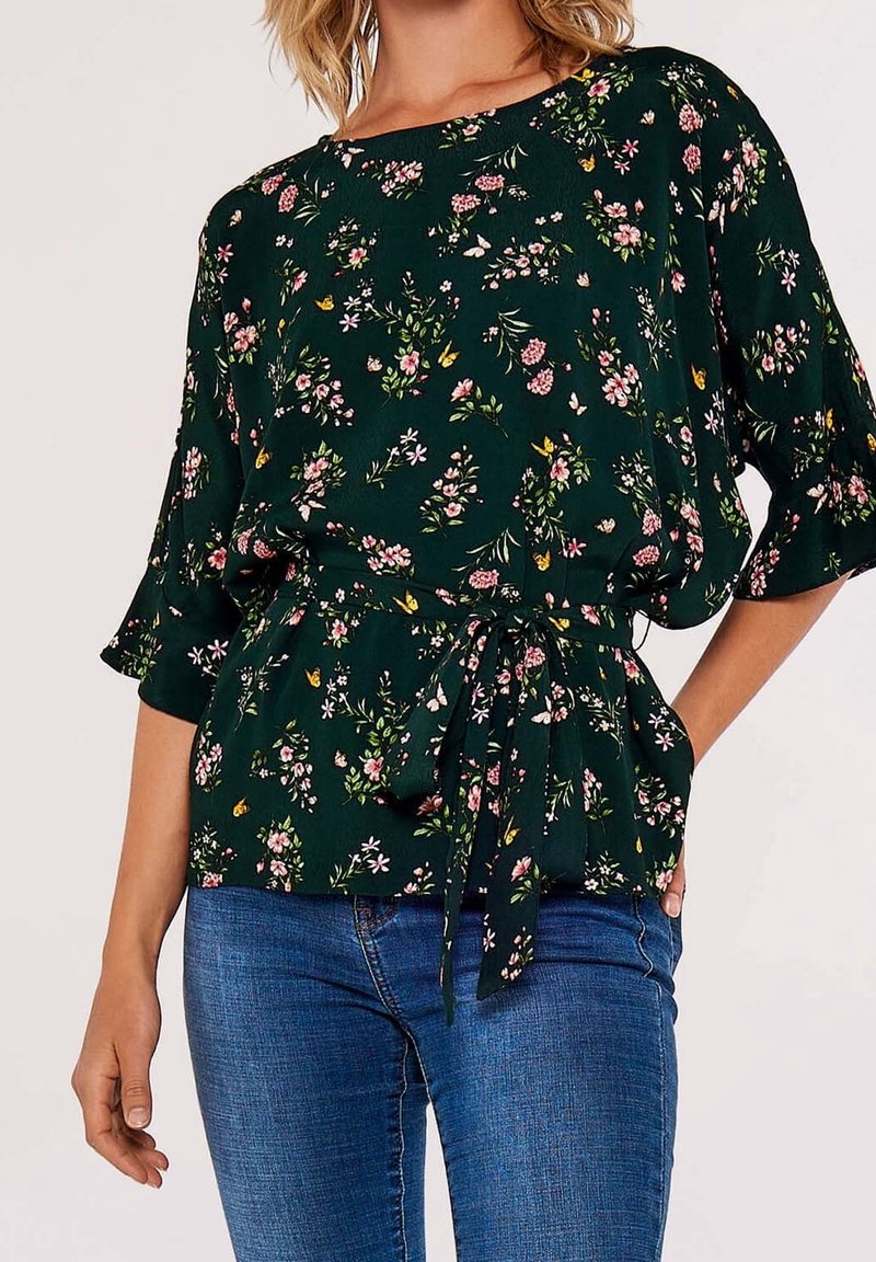 Blouse fleurie vert foncé à manches courtes avec un lien à la taille. Présente un mélange de petites fleurs roses et blanches ainsi que des feuilles.