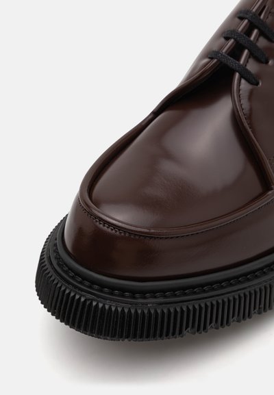 Scarpa in pelle marrone con finitura lucida, punta arrotondata e suola nera con scanalature. Cuciture minime e lacci neri completano il design.