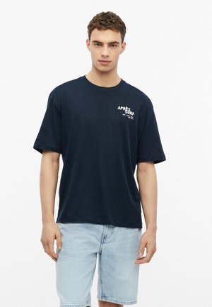 Młody mężczyzna ubrany w granatowy t-shirt "Après Surf" i jasnoniebieskie szorty jeansowe stoi na białym tle.