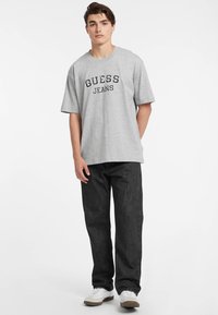 Šviesiai pilkas trumpomis rankovėmis marškinėliai su užrašu "GUESS JEANS" didelėmis juodomis raidėmis. Įvilkta su tamsiais džinsais ir baltais sportbačiais.