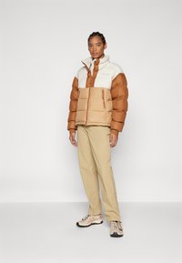 Jachetă puffer maro și crem cu buzunare cu fermoar, asortată cu pantaloni bej și adidași deschiși. Material neted, formă pătrată și guler înalt.