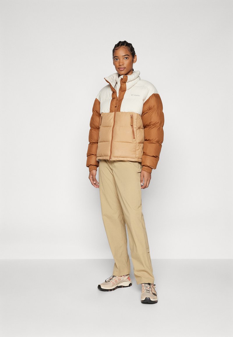 Veste en duvet marron et crème avec des poches zippées, associée à un pantalon beige et des baskets claires. Tissu lisse, forme carrée et col haut.