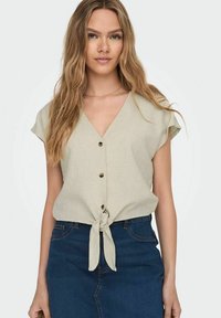 Camicia button-up color beige chiaro con maniche corte e nodo in vita, abbinata a una gonna di jeans. Texture liscia, design casual.