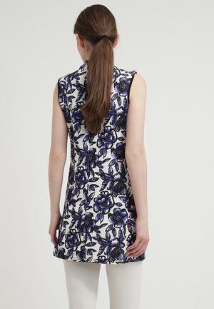 Vrouw met lang haar in een paardenstaart draagt een mouwloze witte tuniek met blauw en zwart bloemmotief en witte broek, gezien van achteren.