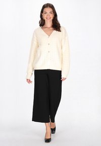 Crème kabelgebreide cardigan met een V-hals, knoopsluiting aan de voorkant en een losse pasvorm, gecombineerd met zwarte hoge taille wijde broek en zwarte hakken.