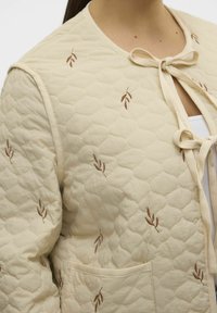 Gequiltede beige jas met bruine bladborduursels. Voorzien van een striksluiting bij de hals en zijzakken. Zacht stof met een gemêleerd patroon.