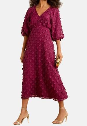 Kobieta w fakturowanej malinowej sukience midi z przezroczystymi bufkami na rękawach, trzymająca małą złotą kopertówkę i nosząca złote sandały na paski.