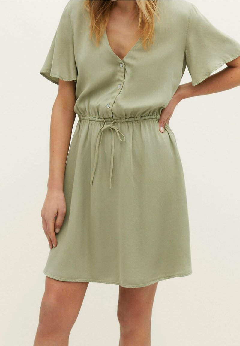 Femme portant une robe vert sauge à manches courtes boutonnée, avec une taille à cordon, debout, une main sur la hanche et l’autre détendue sur le côté.