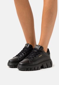 Baskets en cuir noir avec une semelle épaisse, présentant une texture lisse, un bout arrondi et un laçage prononcé. Accents dorés sur la languette et le côté.