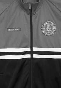 Giacca grigia con strisce bianche e nere, chiusura con zip e toppe ricamate con la scritta "UNFAIR ATHL." e un logo. Finitura testurizzata.