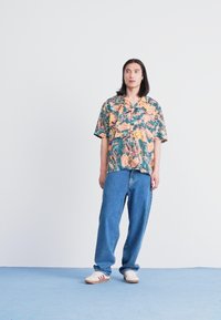 Short-sleeve shirt with a multicolor floral pattern; loose-fit blue jeans. Sneakers with white base and red stripes. Neutral background.