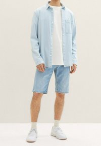 Camisa de mezclilla en azul claro sobre una camiseta blanca, combinada con pantalones cortos en azul claro y zapatillas blancas. La camisa tiene un único bolsillo en el pecho.