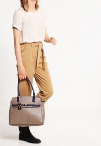 Beige kabelka s námořnickými akcenty, strukturovaný design, dvě horní ucha, přední kapsa na zip a kovový detail s logem.