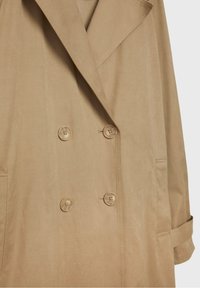 Trench-coat beige à double boutonnage avec quatre boutons assortis, larges revers et brides aux poignets sur un fond uni.