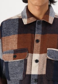 Rutet flanellskjorte i brunt, marineblått og krem; har to brystlommer og svarte knapper. Myk tekstur med klassisk kragedesign.