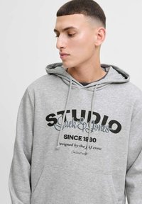 Šedá mikina s kapucí a klokaní kapsou, na přední straně se nachází velký černý a šedý text "STUDIO Jack & Jones" a "od roku 1990".