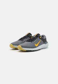 Pelēkie un melnie Nike skriešanas apavi ar dzeltenām zīmogzīmēm un dzelteniem galiem uz baltā fona.