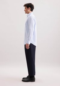 Camicia a maniche lunghe di colore azzurro chiaro con taschino sul petto, abbinata a pantaloni scuri e scarpe nere. L'outfit ha una vestibilità rilassata e una texture liscia.